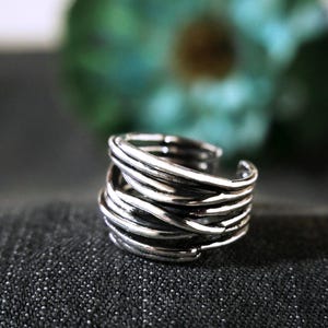 May include: A silver-colored ring with a woven design. The ring features multiple bands of metal intertwined to create a textured appearance. The ring is set against a dark background with a blurred green floral element.
