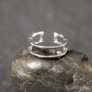 May include: A silver-toned open ring featuring a double-band design with a textured, segmented surface that mimics the appearance of bamboo stalks. The ring is constructed with two parallel bands connected at the sides, creating a hollow centre.