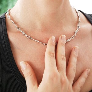 May include: Silver-toned necklace with a barbed wire design. The necklace is worn around the neck and is made of metal. The person is wearing a black shirt.
