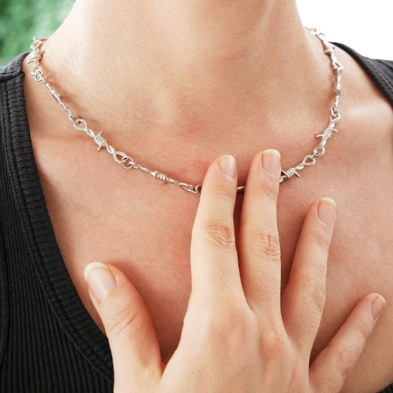 May include: Silver-toned necklace with a barbed wire design. The necklace is worn around the neck and is made of metal. The person is wearing a black shirt.