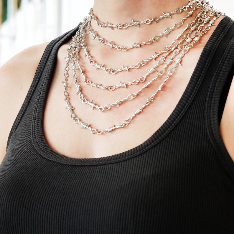 May include: A silver-coloured necklace set featuring multiple strands of barbed wire design. The necklaces are layered, creating a bold statement piece. The jewellery is worn over a black vest top.