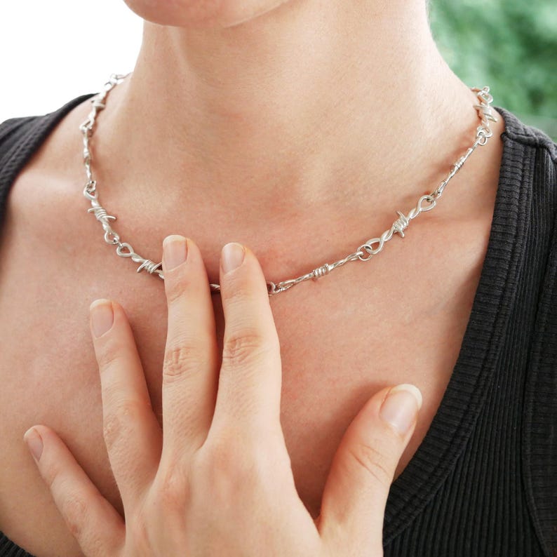 May include: A silver-colored necklace with a barbed wire design. The necklace is worn around the neck and is made of metal. The image shows the necklace on a person wearing a black top.