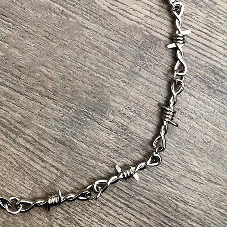 May include: A silver-colored necklace featuring a barbed wire design. The necklace is made of linked metal and has sharp, pointed barbs. The jewelry is displayed on a wooden surface.