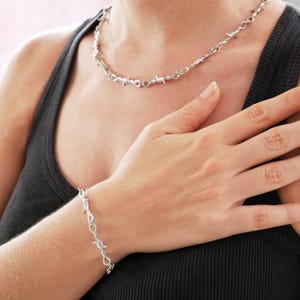 May include: A silver-colored necklace and bracelet set. The jewelry features a barbed wire design. The necklace is around the neck, and the bracelet is on the wrist. The person is wearing a black tank top.