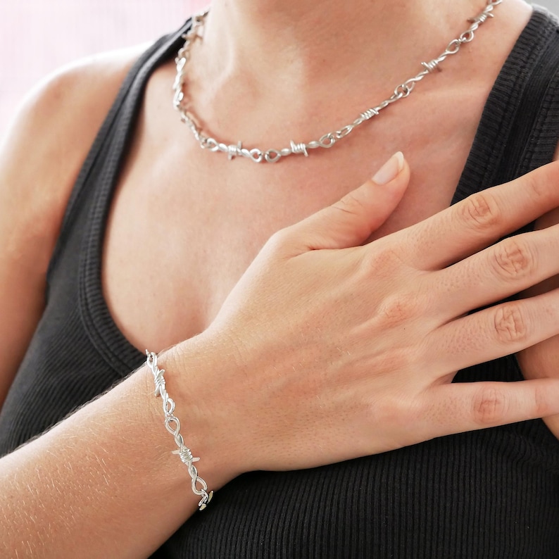 May include: A silver-colored necklace and bracelet set. The jewelry features a barbed wire design. The necklace is around the neck, and the bracelet is on the wrist. The person is wearing a black tank top.