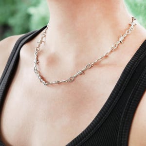 May include: Silver necklace featuring a barbed wire design. The necklace is worn around the neck, resting on a black vest top. The jewellery has a distinctive, edgy style.