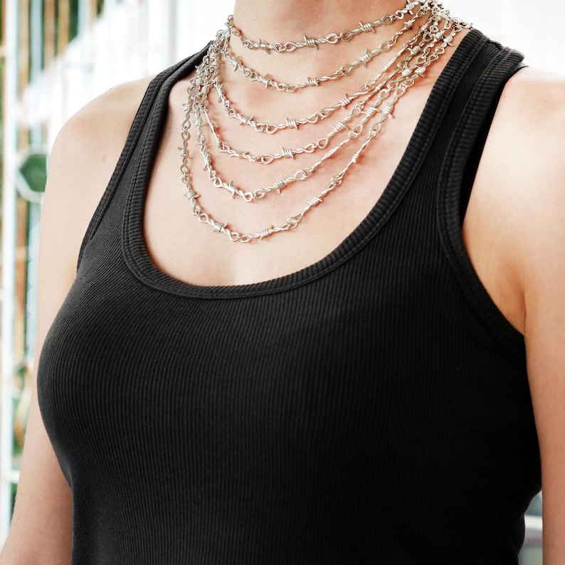 May include: A silver-colored necklace with multiple strands of barbed wire design. The necklace is worn over a black ribbed tank top. The jewelry is a bold statement piece.