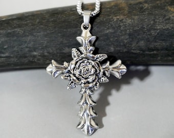Könnte beinhalten: Silberfarbener Kreuzanhänger mit detaillierter Rose in der Mitte. Das Kreuz hat verzierte, spitze Kanten und hängt an einer filigranen Kette. Der Anhänger ist wahrscheinlich aus Metall und wird vor einem neutralen Hintergrund präsentiert.