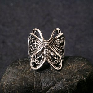 May include: A silver butterfly ring with detailed filigree patterns. The ring showcases openwork wings and a detailed body, giving it a delicate and ornate look. The ring is presented on a dark, textured surface.
