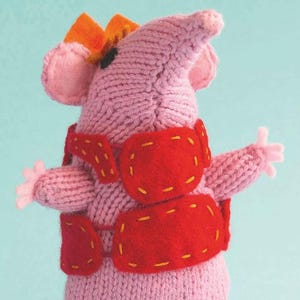 Puede incluir: Un juguete de ratón rosa tejido a mano con un chaleco rojo y una corona naranja. El ratón tiene pies negros y orejas y manos rosas. El chaleco está decorado con costuras amarillas.