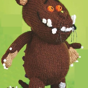 Peut inclure: Un jouet monstre tricoté marron avec des cornes, des dents et des griffes blanches. Le monstre a des yeux orange, un ventre rond et une petite queue. Une version plus petite du monstre est également présente. Le jouet est posé sur un fond vert.