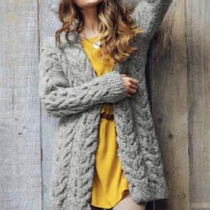 May include: A long, gray cable-knit cardigan. The sweater has a textured pattern and long sleeves. The cardigan is worn over a yellow dress. The background is a wooden wall.