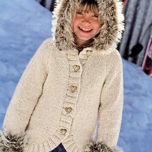 May include: A cream-colored knitted jacket with a faux fur-lined hood and cuffs. The jacket features heart-shaped buttons and a scalloped hem. The background shows a snowy environment, suggesting winter wear.
