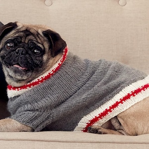 May include: A pug dog wearing a grey knitted sweater with red and white detailing. The dog is lying on a beige surface. The sweater has a ribbed collar and a stripe down the side. The dog is looking at the camera.
