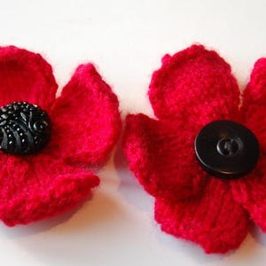 May include: Two handmade red knitted flower embellishments with black button centers. The flowers have five petals each and are displayed against a white background. These decorative flowers can be used for various craft projects.