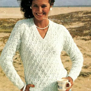 Peut inclure: Un pull en tricot texturé bleu clair avec un col en V. Le pull à manches longues présente un motif en losanges et est porté avec un bracelet blanc et un collier de perles. Une tasse avec un motif floral est tenue à la main.