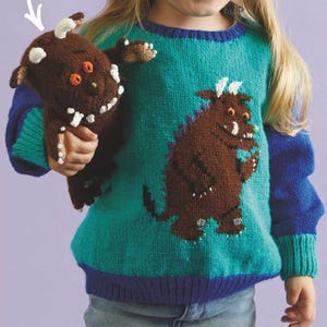 Könnte beinhalten: Ein teal- und blau gestrickter Pullover mit einem braunen Monster-Design. Der Pullover hat blaue Ärmel und eine blau-violette Bordüre. Ein passendes braunes Monster-Plüschtier wird im Bild gehalten.