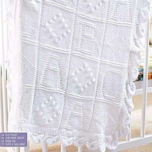 May include: A white knitted baby blanket with a scalloped edge, displayed in a crib. The blanket features a grid pattern with textured squares, some containing letters and diamond shapes. The text in the image says "Easy Peasy", "A bit more tricky", "Hard-ish", and "Quite a challenge".