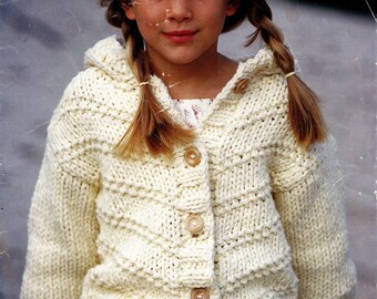 Könnte beinhalten: Cremefarbene Kapuzenstrickjacke mit Holzknöpfen. Der Strickpullover hat ein strukturiertes Muster und wird von einer Person mit blonden Zöpfen getragen. Die Texte "DENIM ULTRA" und "SIRDAR" sind sichtbar.