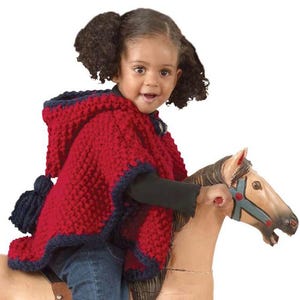 May include: A child's red knitted poncho with navy blue trim and hood. The poncho features a textured pattern and tassel. The child is seated on a wooden rocking horse with a painted face and blue bridle.