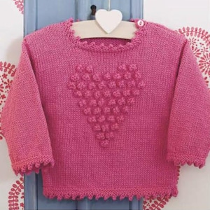 May include: A pink knitted sweater with a heart-shaped design on the front. The sweater has long sleeves and a scalloped edge around the neckline, cuffs, and hem. A small button is visible at the shoulder.