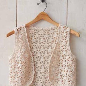 May include: A cream-colored, open-front crochet vest hangs on a wooden hanger. The vest features a delicate, openwork pattern and a sleeveless design. The garment is displayed against a light-colored wooden background.
