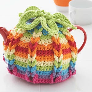 Pode incluir: Uma chaleira vermelha com uma colorida capa de chá de crochê. A capa apresenta um arco-íris de cores, incluindo rosa, azul, verde, laranja e amarelo, com um design verde em forma de folha no topo. A capa é intrincadamente trançada.