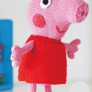 May include: A knitted pink pig doll wearing a red dress. The pig has a large snout, small ears, and black eyes. The doll's arms and legs are also pink, and it has black shoes. The doll is a children's toy.