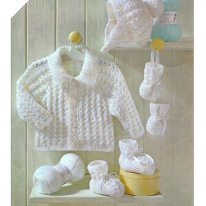 Puede incluir: Un conjunto de ropa de bebé de punto blanco, que incluye una chaqueta, un gorro y patucos, expuestos en una estantería. El conjunto incluye dos ovillos de lana y está diseñado para tallas prematuras. La marca "Sirdar" es visible.