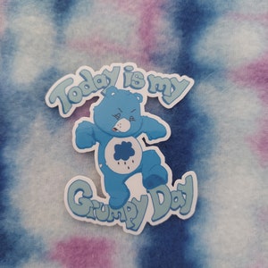 May include: A blue Care Bear with a grumpy expression and a rain cloud on its chest. The bear is standing with its arms crossed and its feet together. The text "Today is my Grumpy Day" is written in a blue and green font around the bear.