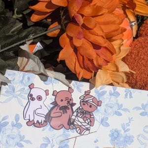May include: A light blue and white floral patterned card featuring three cartoon bears dressed in Halloween costumes: one as a ghost, one as a vampire with a cape, and one as a mummy wrapped in bandages. The word Twenty is written in gold script.