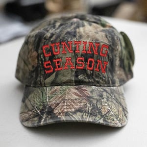 May include: Camouflage baseball cap with the words "HUNTING SEASON" embroidered in red. The cap has a curved peak and a classic six-panel design, suitable for outdoor pursuits.