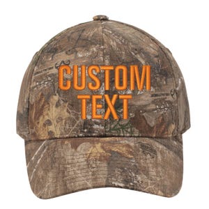 May include: A brown and tan camouflage baseball cap with an orange embroidered "CUSTOM TEXT" on the front.