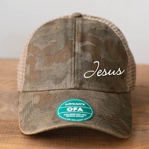 May include: A camouflage trucker hat with a white embroidered "Jesus" on the front. The hat has a brown mesh back and a green Legacy OFA label on the side.