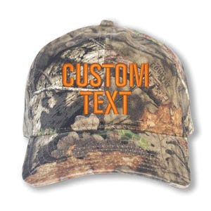 May include: A camouflage baseball cap with the words "CUSTOM TEXT" embroidered in orange thread across the front. The cap features a curved brim and a structured crown, suitable for outdoor activities.