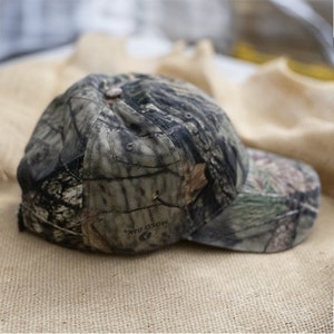 May include: A camouflage baseball cap with a curved brim. The cap features a pattern of greens, browns, and blacks, with the words "Mossy Oak" visible on the side. The cap is resting on a tan, textured surface.