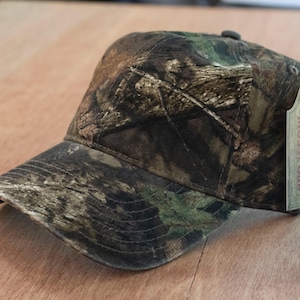 May include: A baseball cap featuring a brown, green, and black camouflage pattern. The cap has a curved brim and a structured crown. Attached to the side is a rectangular cardboard tag with the text MOSSY OAK and a tree logo printed on it.