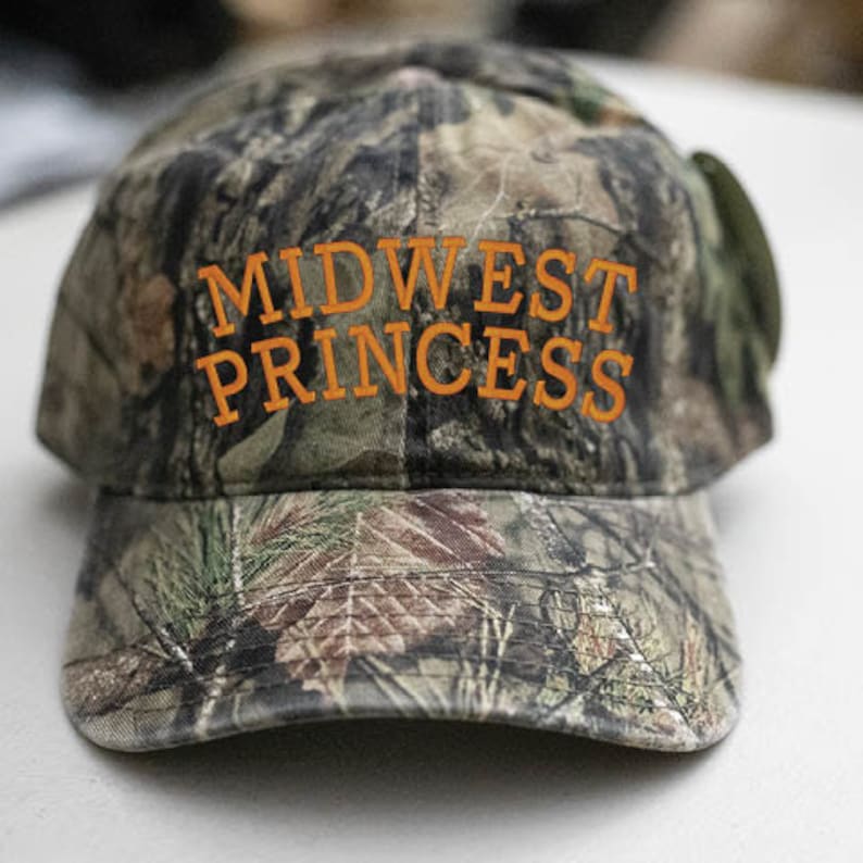 May include: A camouflage baseball cap with the text "MIDWEST PRINCESS" embroidered in orange.