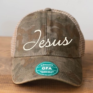 Może przedstawiać: Kamuflażowa czapka bejsbolowa z napisem "Jesus" wyhaftowanym białą czcionką. Czapka ma siatkowy tył i zielono-białą metkę z napisem "Legacy OFA". Kapelusz jest wykonany z materiałów pochodzących z recyklingu.