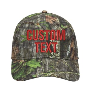 May include: A camouflage baseball cap with the words "CUSTOM TEXT" embroidered in red. The cap features a curved brim and a button on top. The pattern includes shades of green, brown, and tan.