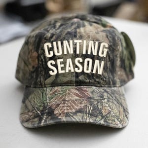 May include: Camouflage baseball cap with the words "CUNTING SEASON" in white block letters. The cap has a standard peak and a classic six-panel design. The camouflage pattern includes shades of green, brown, and tan.