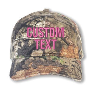 May include: A camouflage baseball cap with the words "CUSTOM TEXT" embroidered in pink. The cap features a curved brim and a classic six-panel design, perfect for outdoor activities or casual wear.