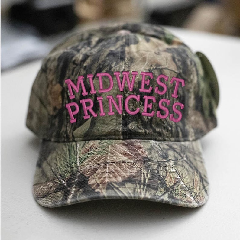 May include: Camouflage baseball cap with the text "MIDWEST PRINCESS" embroidered in pink.