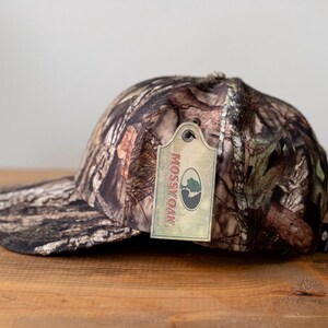 May include: A camouflage baseball cap with a curved brim. The cap features a brown, green, and tan pattern. A tag with the "Mossy Oak" logo is attached to the cap.