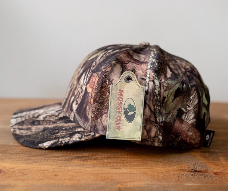 May include: A camouflage baseball cap with a curved brim. The cap features a brown, green, and tan pattern. A tag with the "Mossy Oak" logo is attached to the cap.