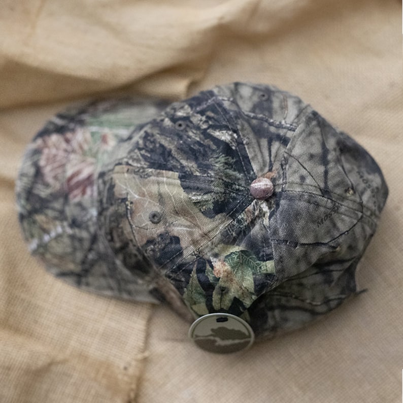 May include: Camouflage baseball cap in shades of green, brown, and tan. The cap features a curved brim and a button on top. A small, round tag with a nature scene hangs from the cap.