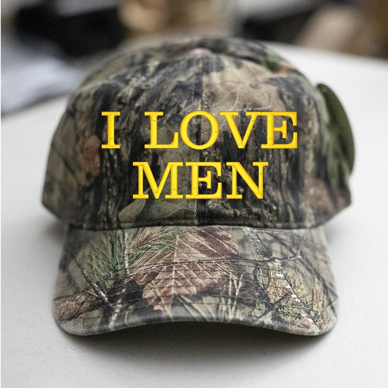 Op de afbeelding: Een camouflage baseballpet met de tekst "I LOVE MEN" geborduurd in geel.