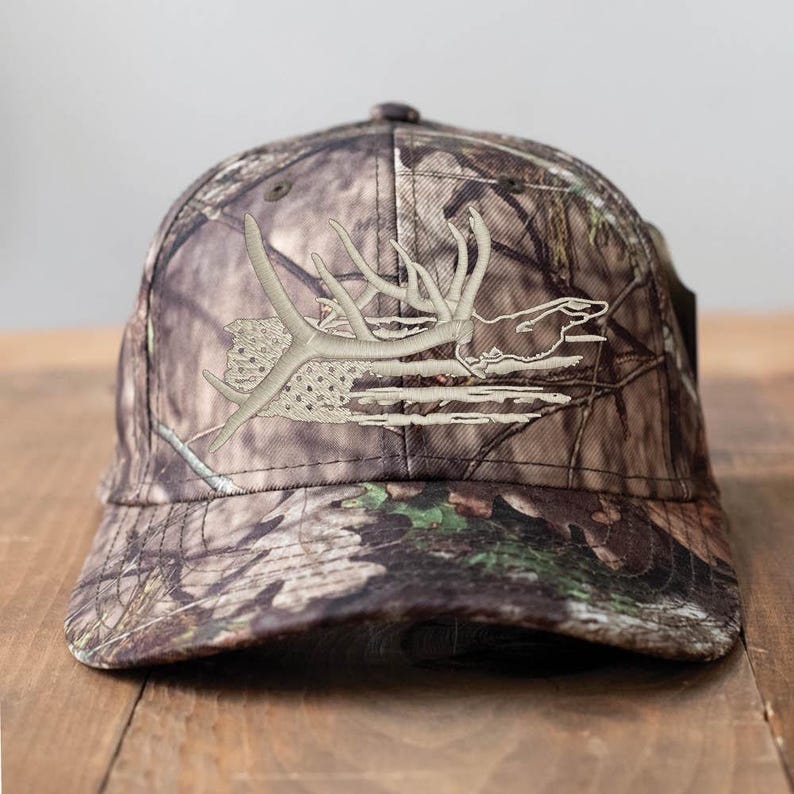 May include: Camouflage baseball cap with an embroidered antler design in shades of beige and tan. The cap features a curved brim and a classic six-panel construction, suitable for outdoor activities.