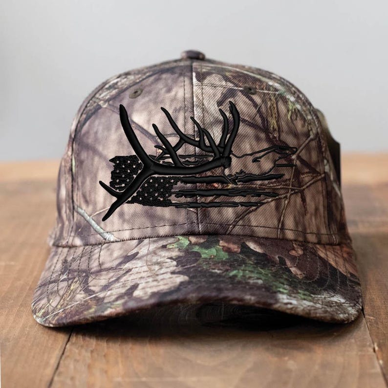 May include: Camouflage baseball cap featuring a black embroidered design of deer antlers and a distressed American flag. The cap has a curved brim and adjustable strap, perfect for outdoor activities or casual wear.
