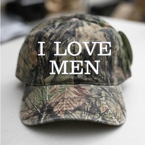 Op de afbeelding: Een camouflage baseballpet met de tekst "I LOVE MEN" in witte letters.
